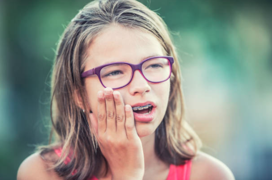 Girl with glasses and braces holding her mouth in pain
